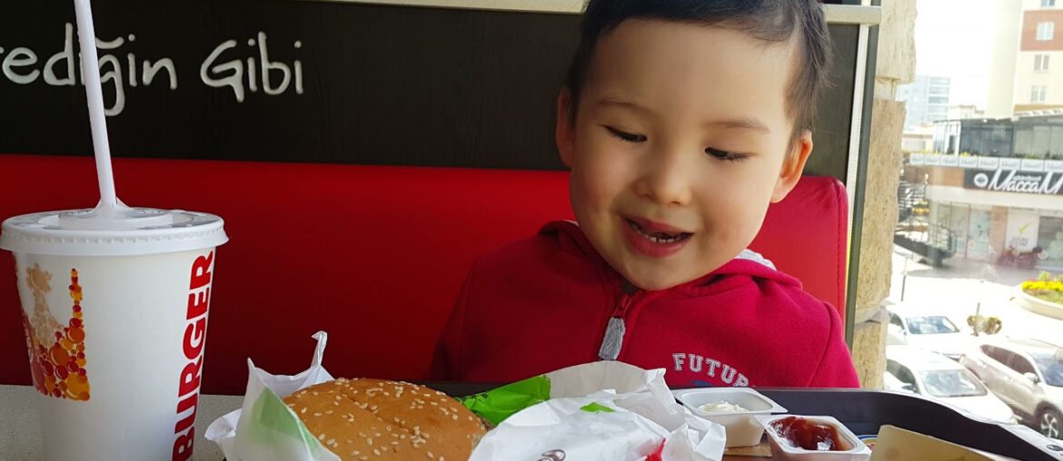 Sweet baby boy eating hamburger with ketchup, fries and juice in Burger King fast food restaurant.