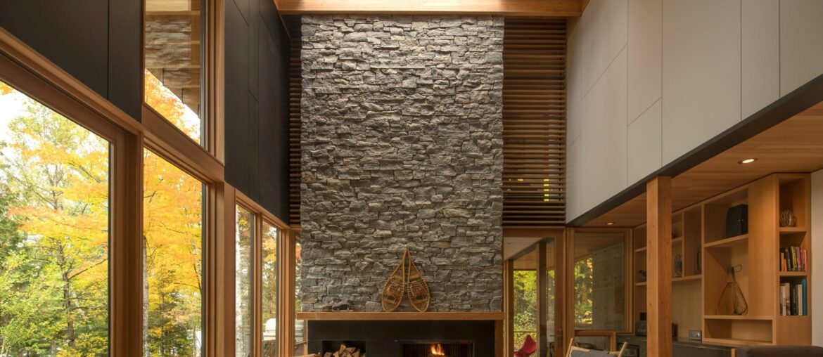 Tall wood living room with a fireplace clad with locally sourced granite in a woodland retreat located along the shores of Contau Lake, Haliburton County, Ontario, Canada [2364×2432]