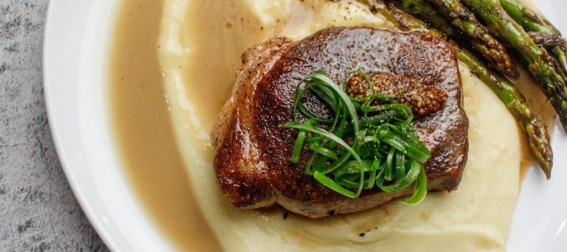 Dry aged pork chop on potato purée served with pork stock reduction and broiled asparagus, topped with sweet pickled mustard seeds and scallions
