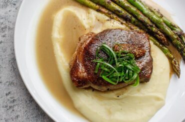 Dry aged pork chop on potato purée served with pork stock reduction and broiled asparagus, topped with sweet pickled mustard seeds and scallions